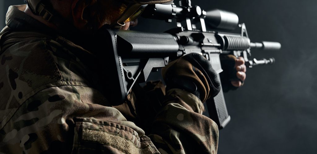 Side close up of old military officer holding sniper rifle and aiming. Crop of bearded veteran in camouflage uniform and helmet posing with gun in smoky dark atmosphere. Concept of military.